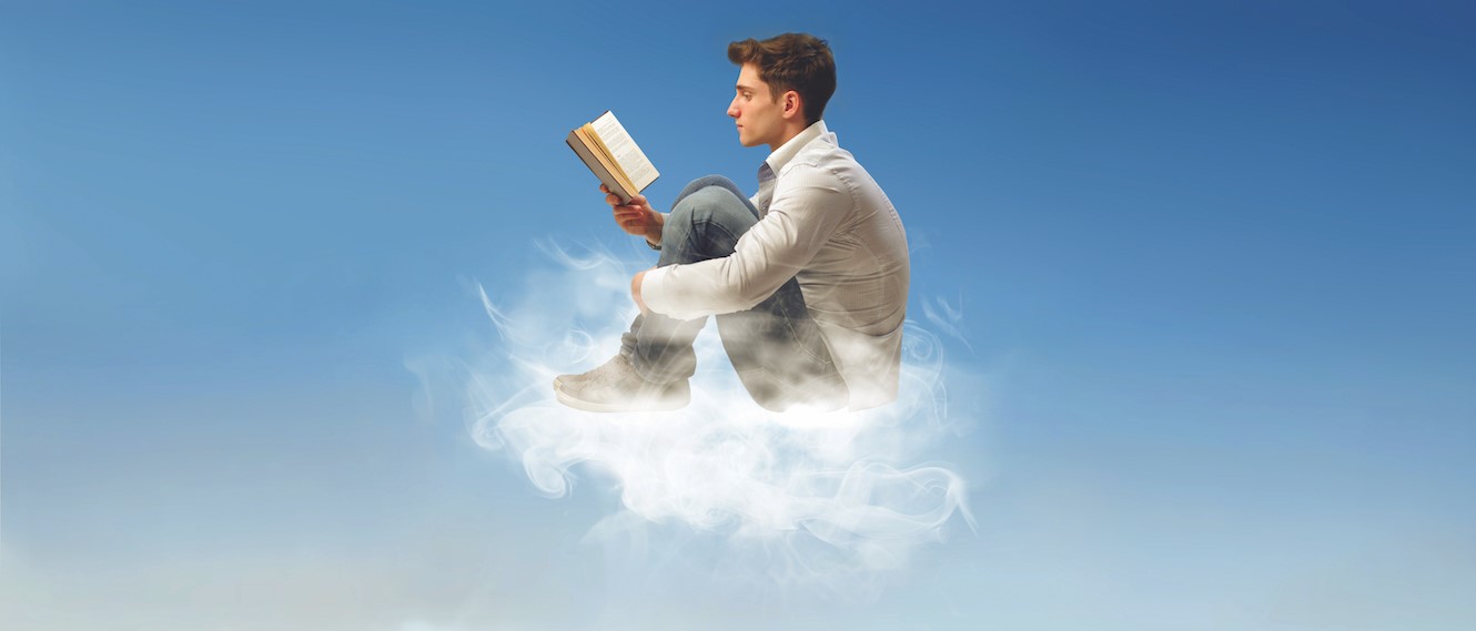teenage boy sits on a wispy white cloud in the blue sky reading a book