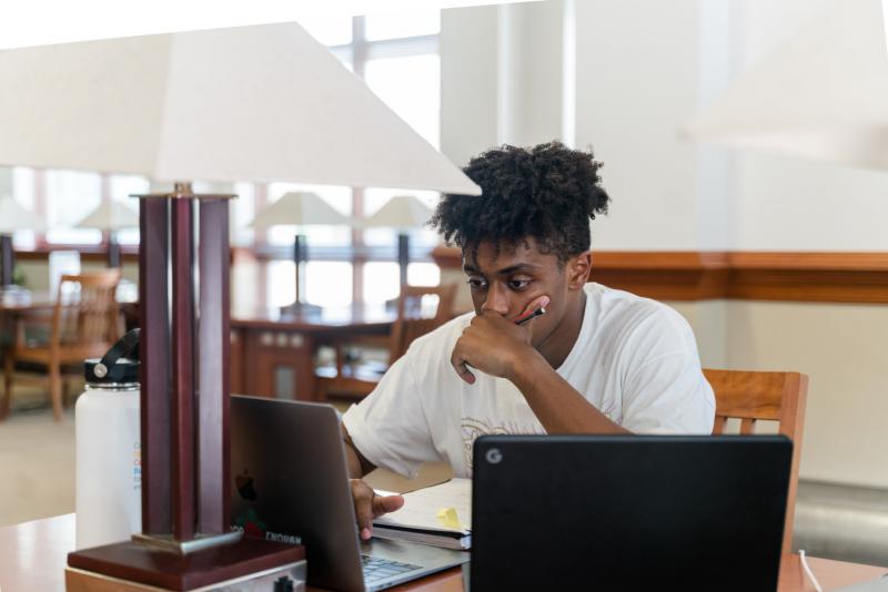 a young man sits at a library desk with two laptops open in front of him and a hand held pensively to his chin