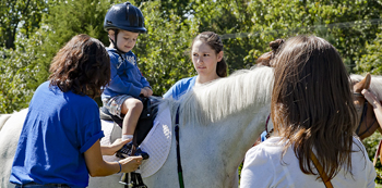 Two trainers help secure a young boy atop a horse while boy's mother looks on