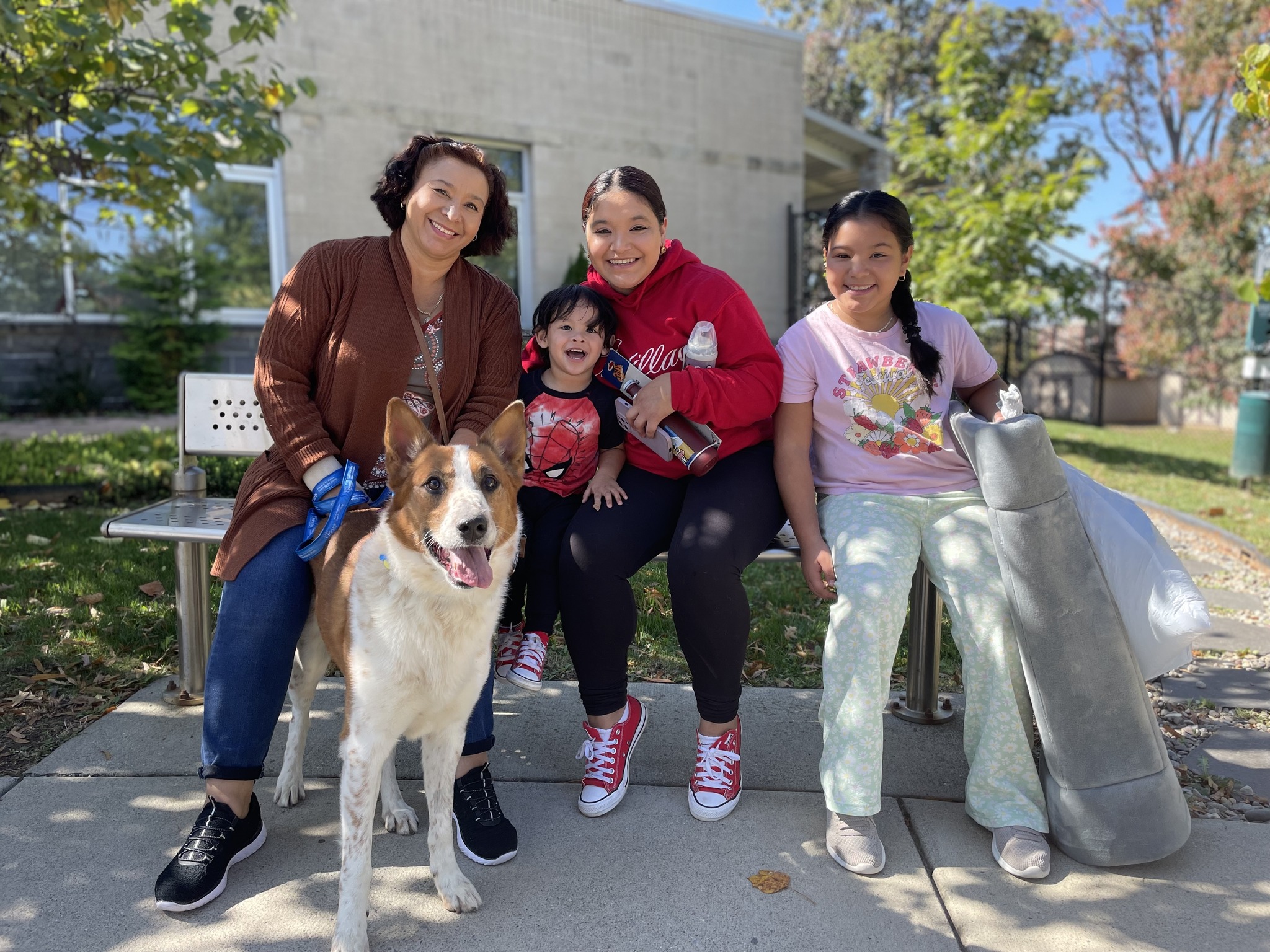 A smiling family of four sits on a bench in the tree-dappled sunshine. They have just been reunited with their beloved dog. Everyone, including their dog, is overjoyed!