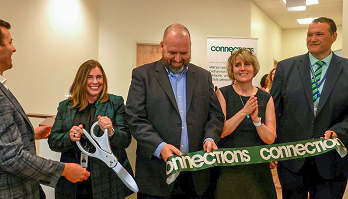 A group of smiling individuals in business dress cut a large ceremonial ribbon with the word “Connections” on it.