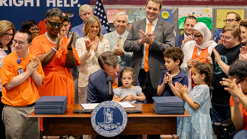 Virginia Governor Glen Youngkin sits at a desk signing a stack of official documents in a ceremony with a young girl sitting on his lap and a diverse group standing around him applauding.
