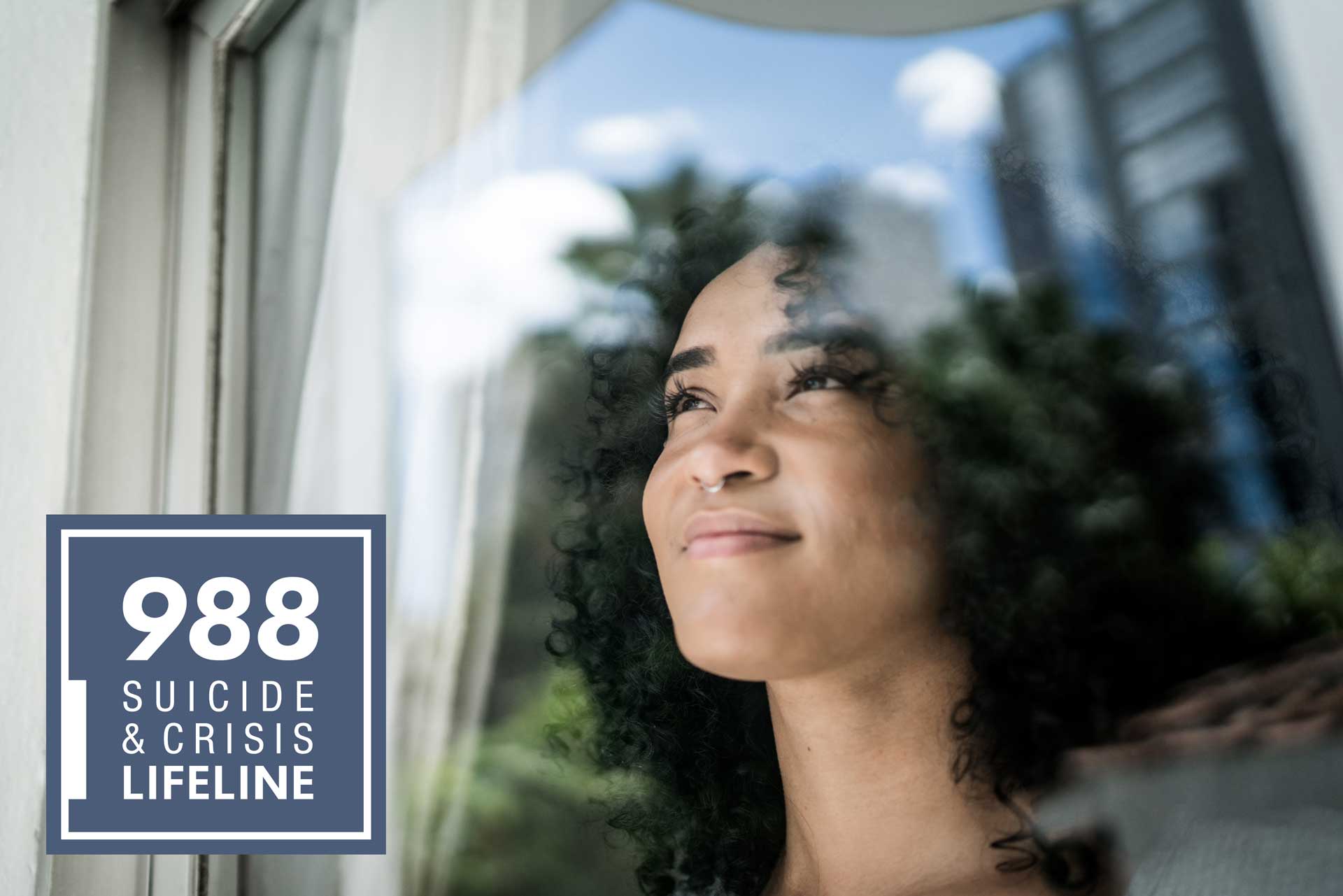 woman looking out a window with hopeful eyes and 988 suicide and crisis lifeline logo overlaid in bottom left of image