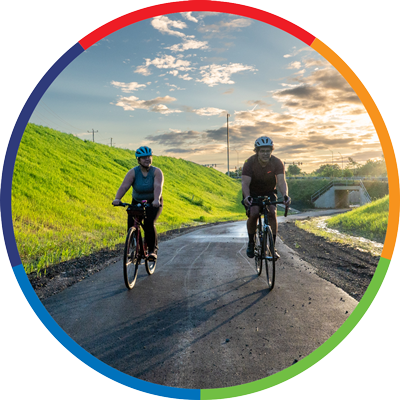 Two individuals ride bikes on a path with a green hill and sun and clouds in the background.