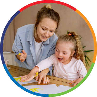 a woman in a blue shirt assists a smiling child as she goes about hand painting on paper