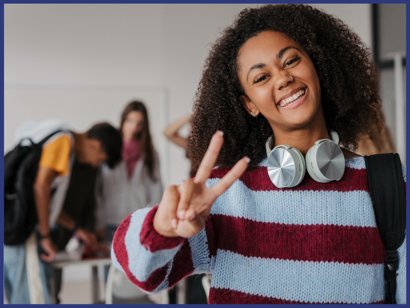 A girl with headphones around her neck and wearing a sweater holds up a peace sign and smiles.