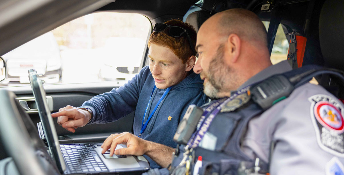 A police officer and a behavioral health professional look at a computer while sitting in a police car.
