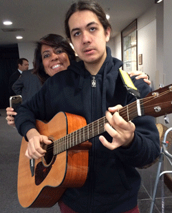 Photo of young man playing guitar, with his mother behind him