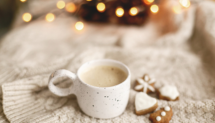 a white mug with coffee rests on a white blanket with gingerbread cookies next to it and holiday lights in the background