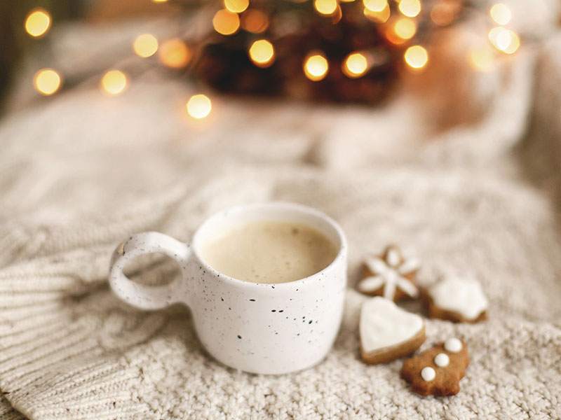a white mug with coffee rests on a white blanket with gingerbread cookies next to it and holiday lights in the background