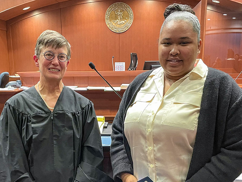 A woman in judicial robes stands next to a woman in professional attire in a courtroom.