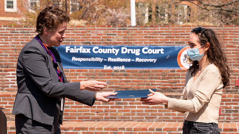 A woman in a suit hands a certificate to another woman in a courthouse courtyard.