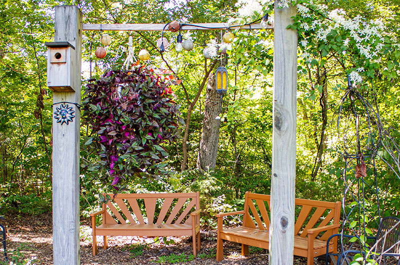 A wooden gate, adorned with flowers and hanging lights, leads to a pair of wooden park benches in a green garden.