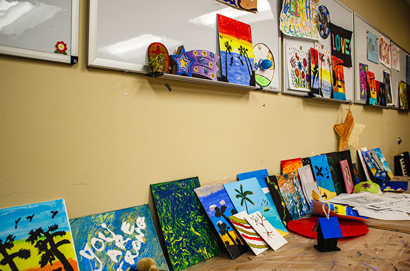 Paintings and handicrafts sit on a table along a wall in an art room.