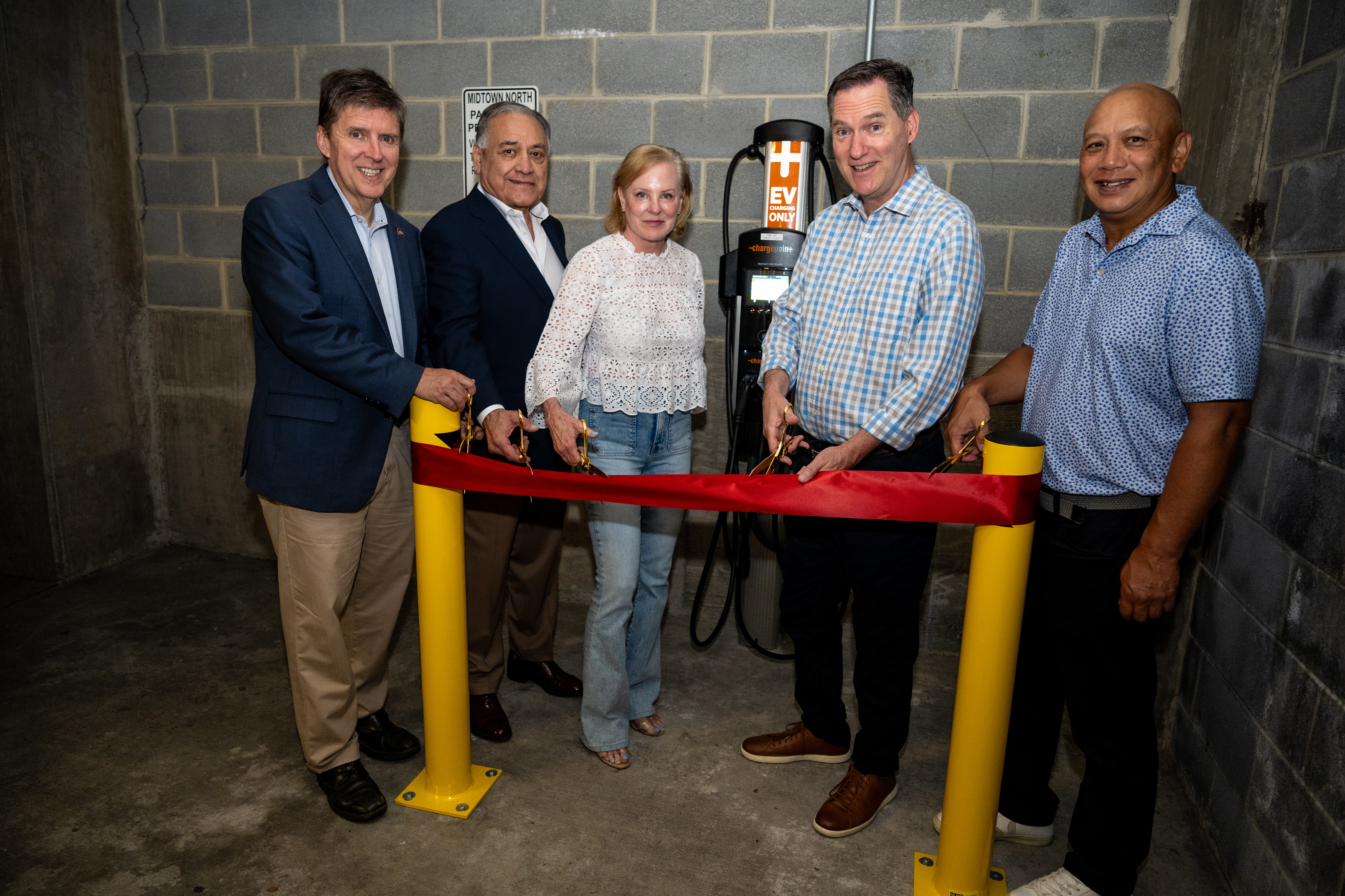  Five individuals stand in a parking garage cutting a red ribbon in front of a newly installed electric vehicle charging station