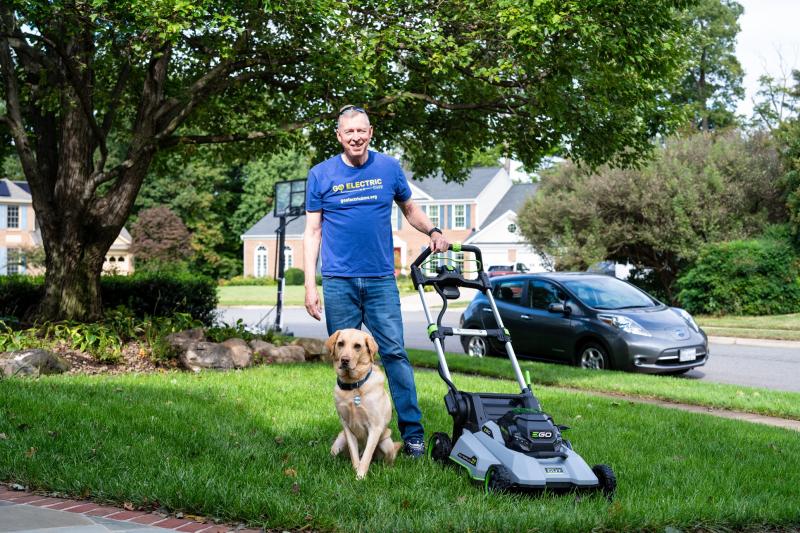A person standing on a lawn with a dog and an electric lawn mower in a suburban neighborhood.