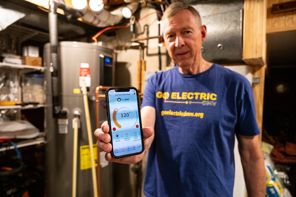 A person in a basement holding a smartphone showing a water heater control app.