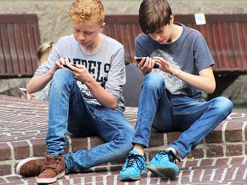 two teenagers sitting on curb using cell phones