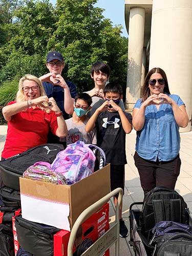 Touching Hearts backpack drive six people standing with backpacks forming hearts with hands