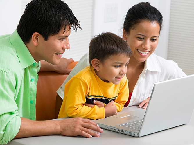 two adults and toddler sitting at table looking at laptop