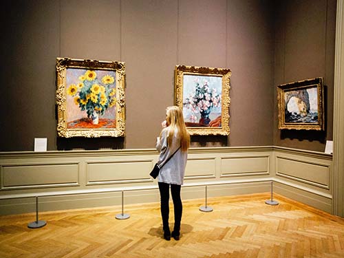 person standing and looking at three paintings in museum