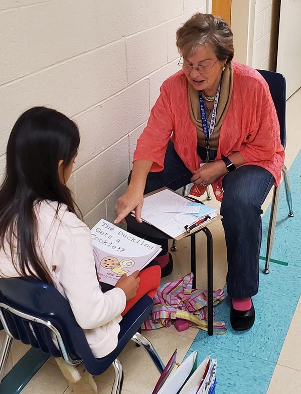 Photo of an older woman tutoring a grade-school child in a school hallway.