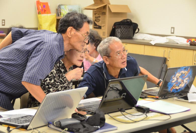 Photo of three adults looking at a laptop screen during a computer tutoring session. 