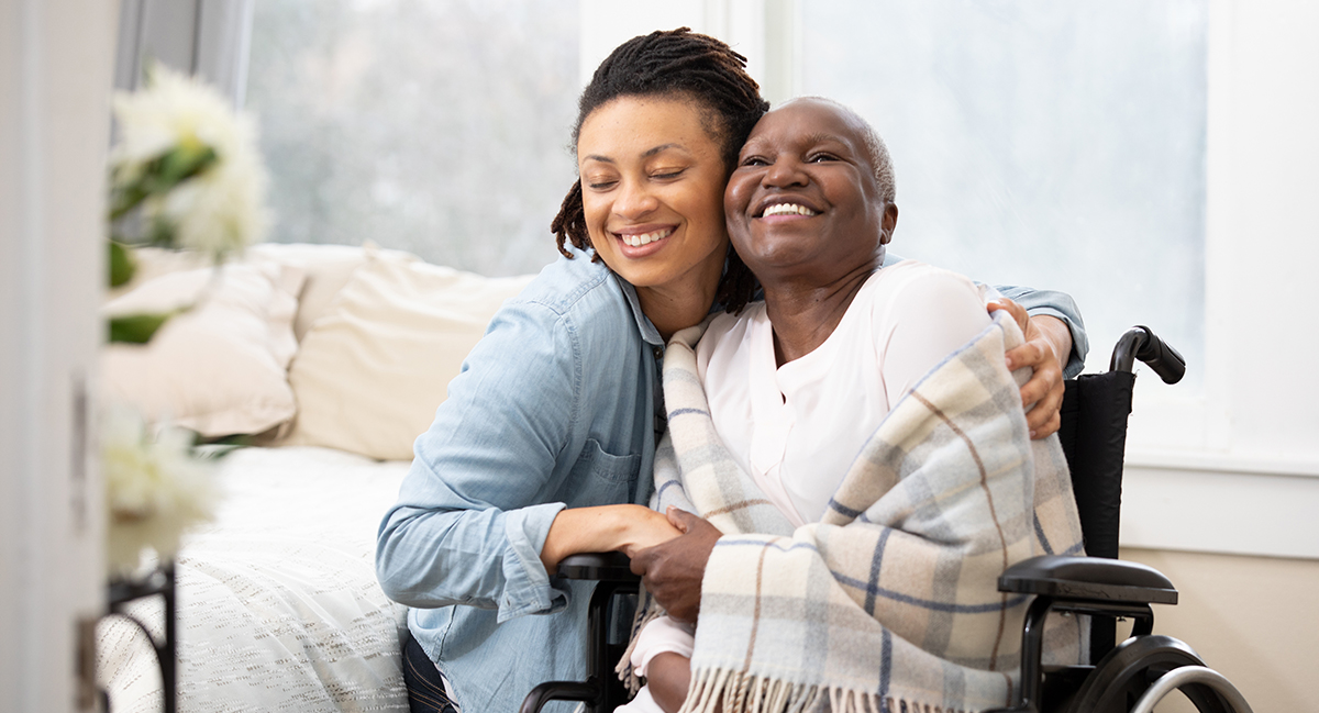 Photo of a woman hugging another woman who is seated in a wheelchair with a blanket wrapped around her.
