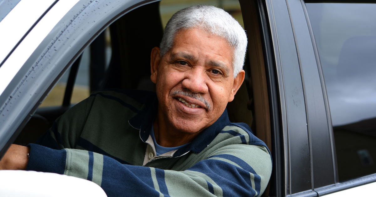 Photo of an older man sitting in the driver seat of a car, looking out the window.