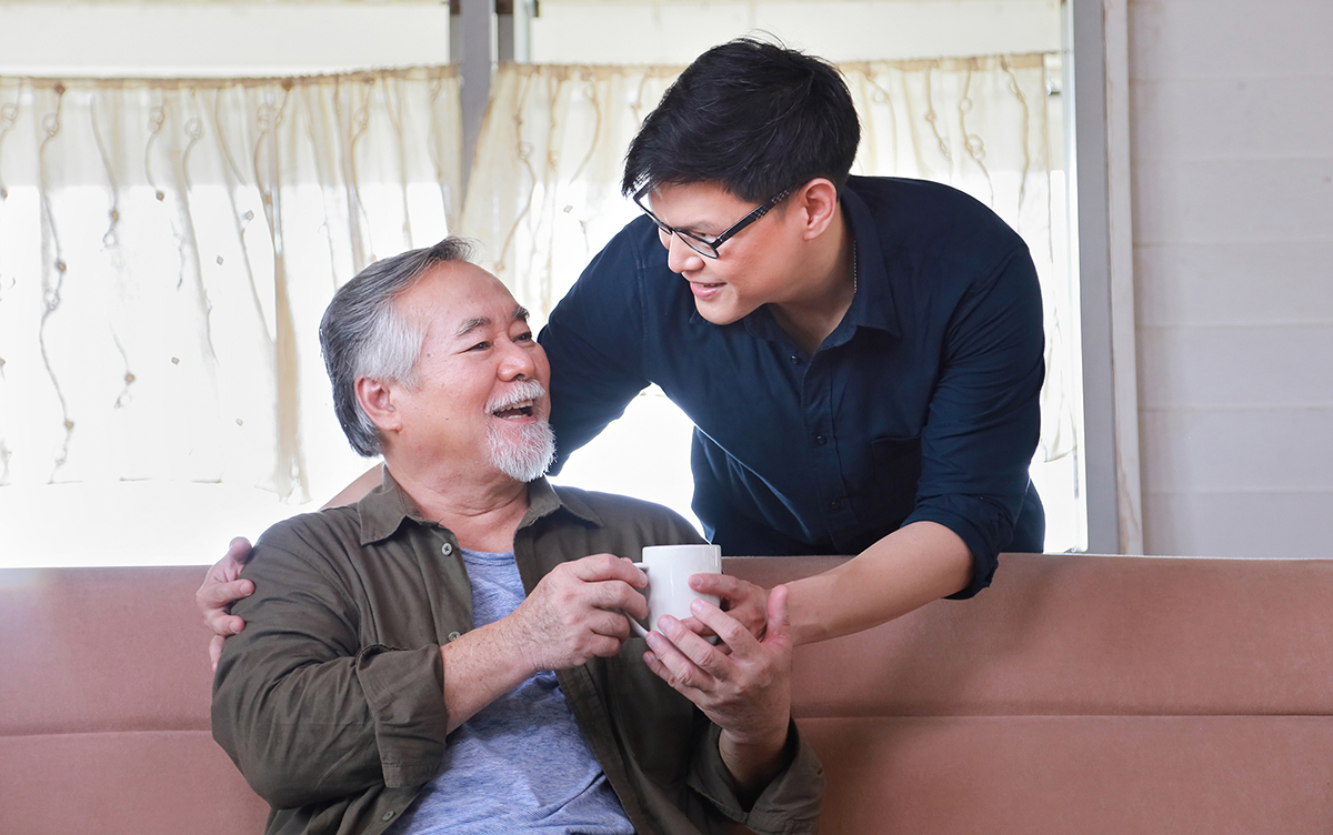 Photo of a young Asian man handing a beverage to a seated older Asian man.