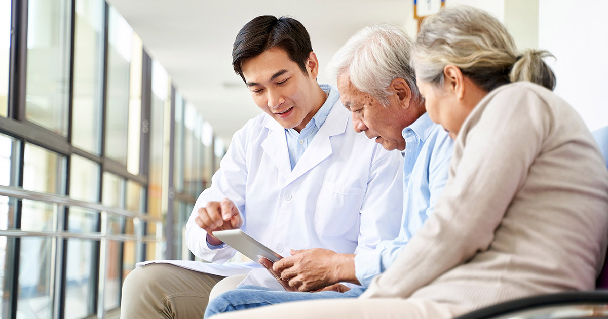 Photo of a young Asian doctor talking to an older couple while showing them something on a tablet device. 