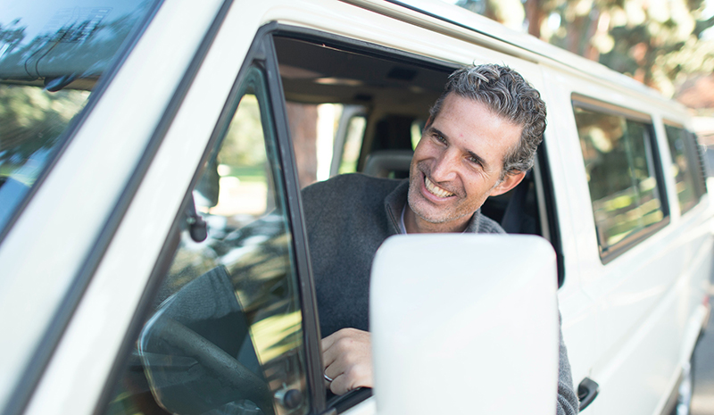 Photo of a smiling man sticking his head out of the window of a van as he drives.