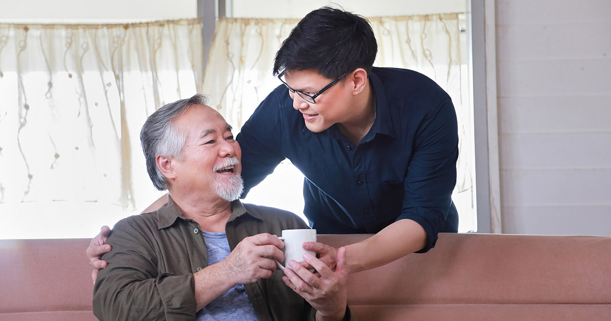 Photo of a young Asian man handing a beverage to a seated older Asian man.