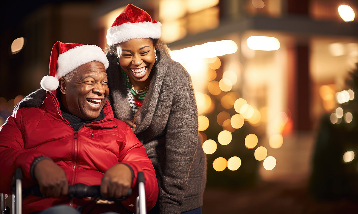 Photo of an older man in a wheelchair and a younger woman laughing together while wearing Santa hats.
