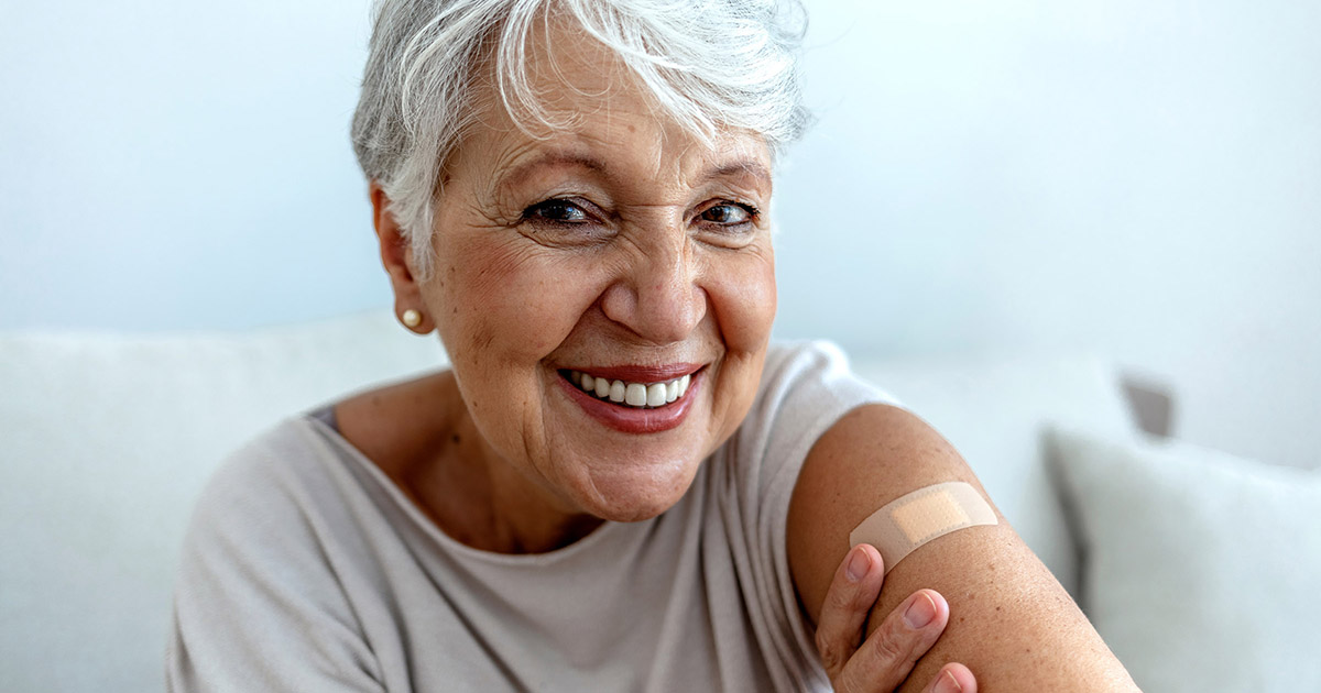 An older woman smiles while she shows of her arm, where a band-aid is applied to her shoulder.