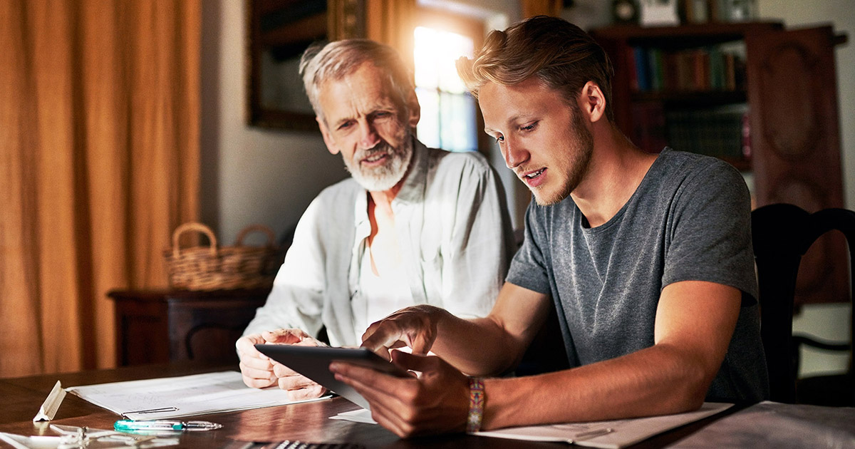 A younger man points to something on an iPad while sitting at a table with an older man.