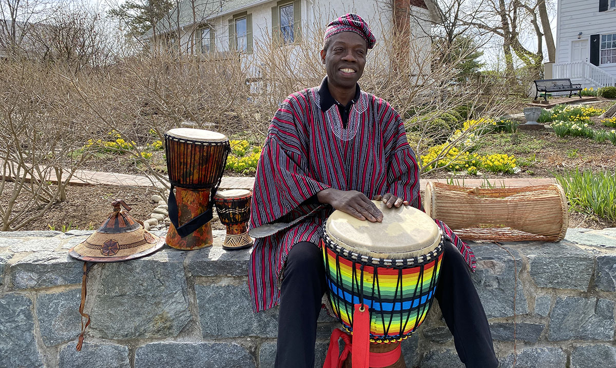 Photo of Kofi Dennis playing a djembe drum outdoors in a park with two smaller djembe drums sitting nearby.