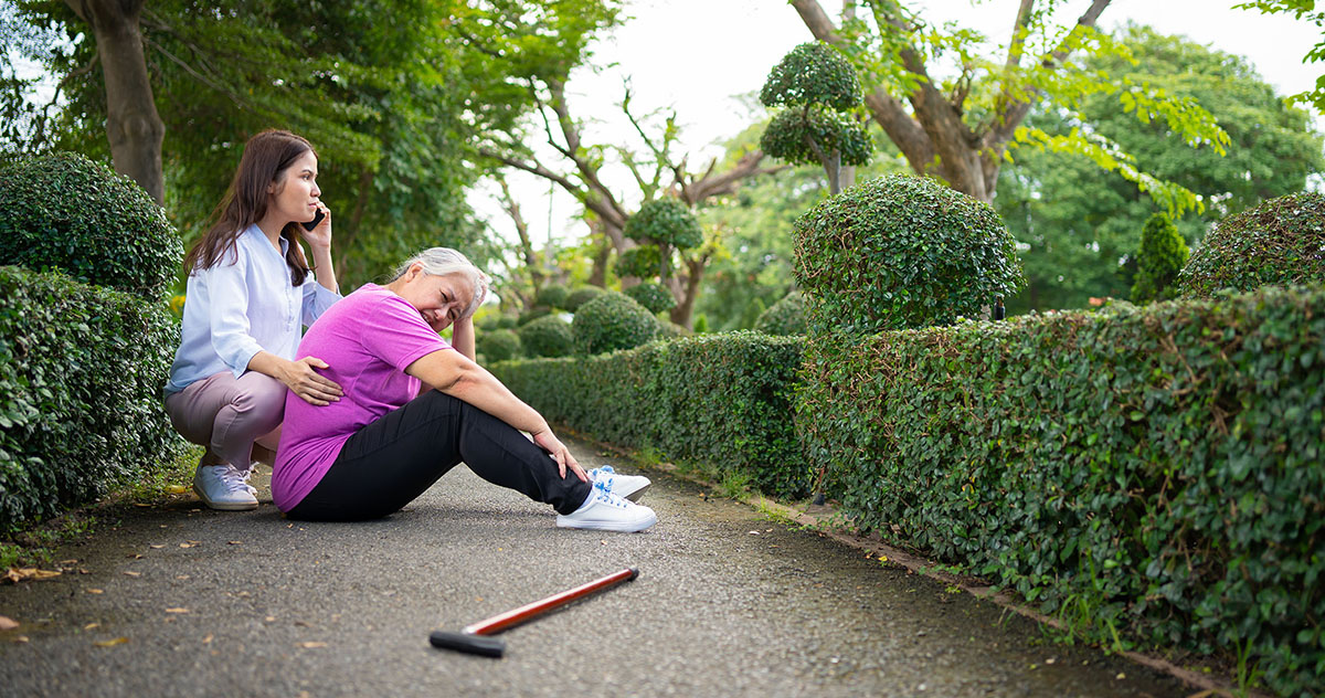 Photo of an older woman sitting on the ground clutching her head while a younger woman makes a call using a smartphone.