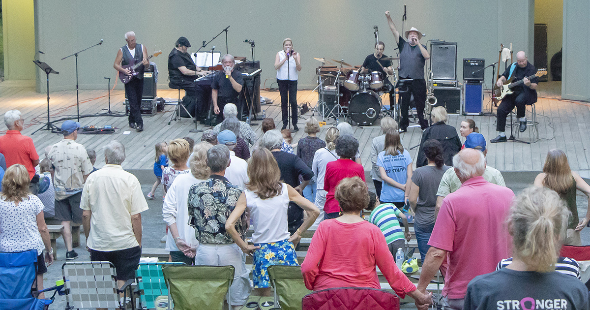 Photo of a band performing on stage at an outdoor concert with audience members seated in lawn chairs and standing to watch the performance.