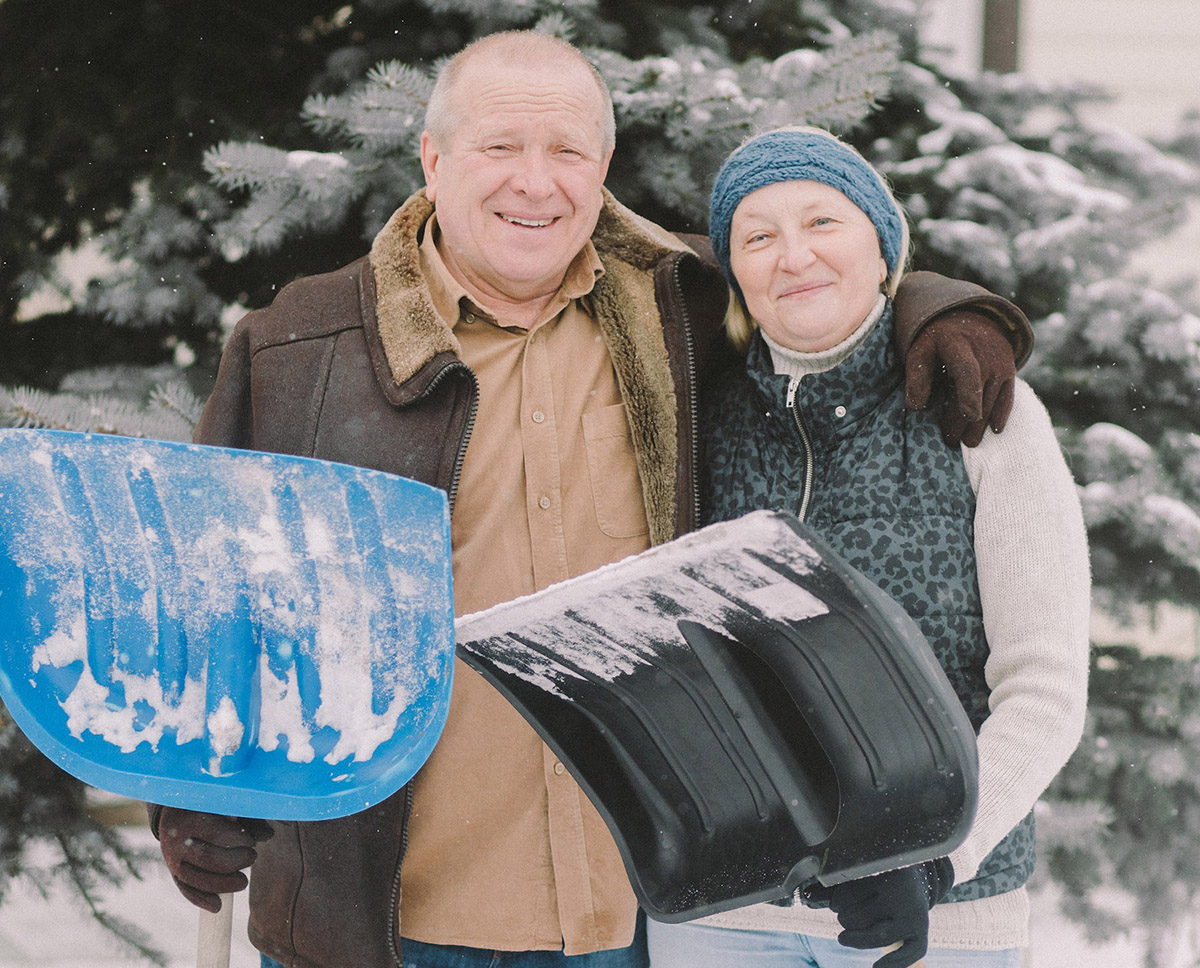 An older couple standing together outdoors in the snow, smiling while holding snow shovels.