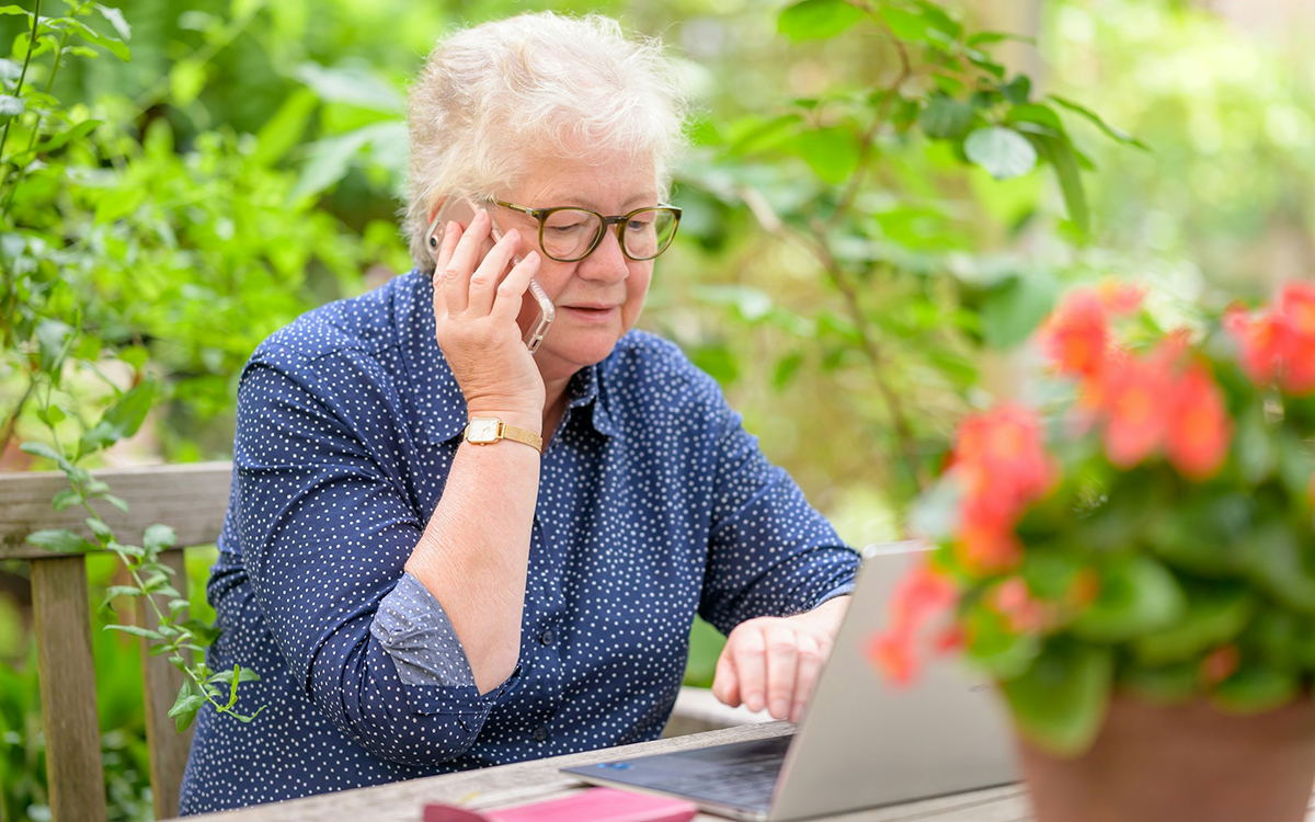 Photo of an older woman seated outdoors talking on a cell phone while seated at a laptop with a concerned look on her face.