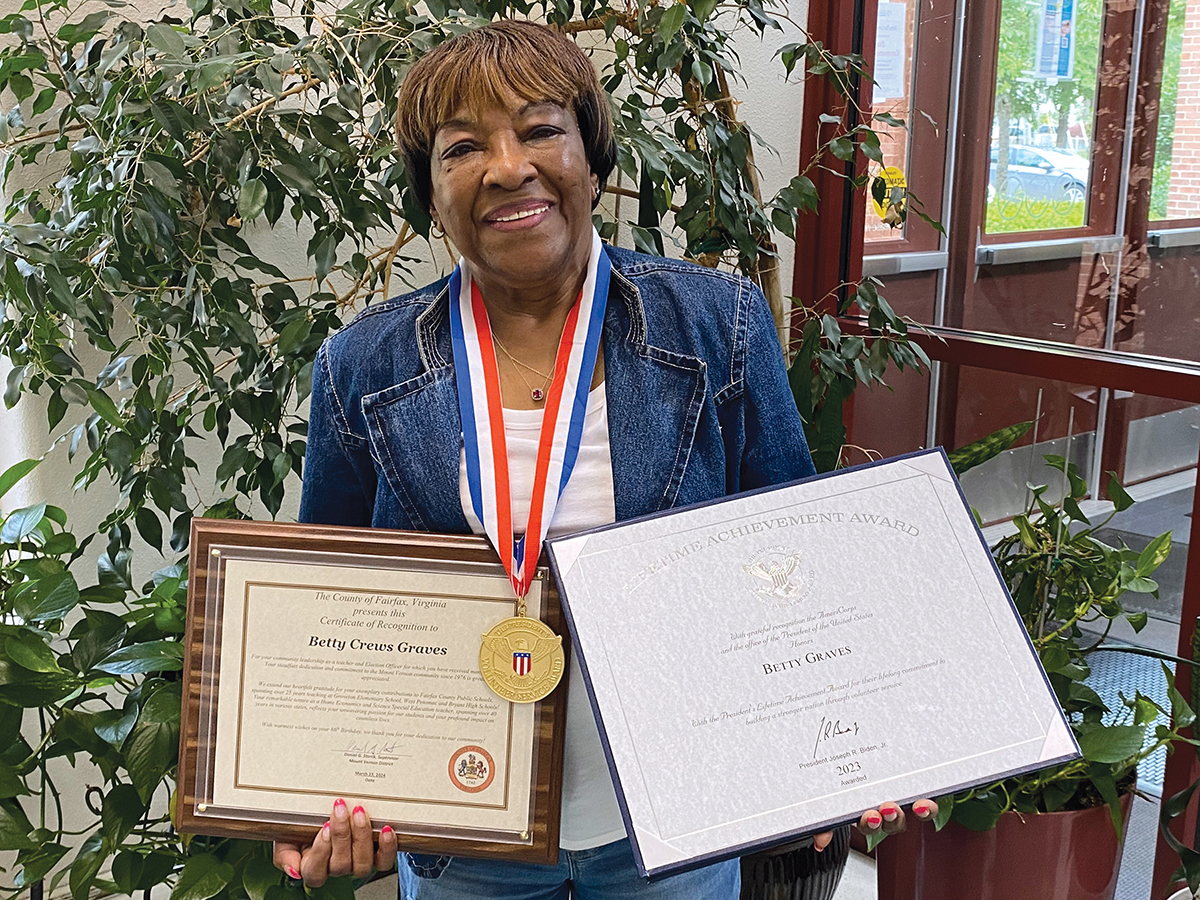 Photo of Betty Graves smiling while holding up two plaques she recently received to recognize her volunteer work. 