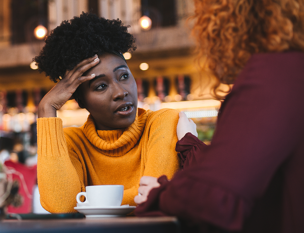 Photo of a stressed-out looking woman talking to a friend while seated in a coffee shop. 