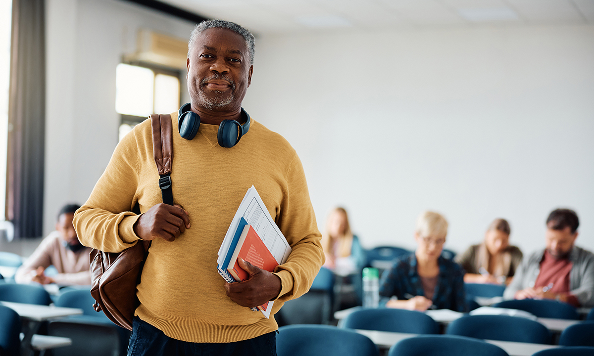 Photo of an older man with a backpack slung over his shoulder standing in a classroom with other adult students seated behind him.