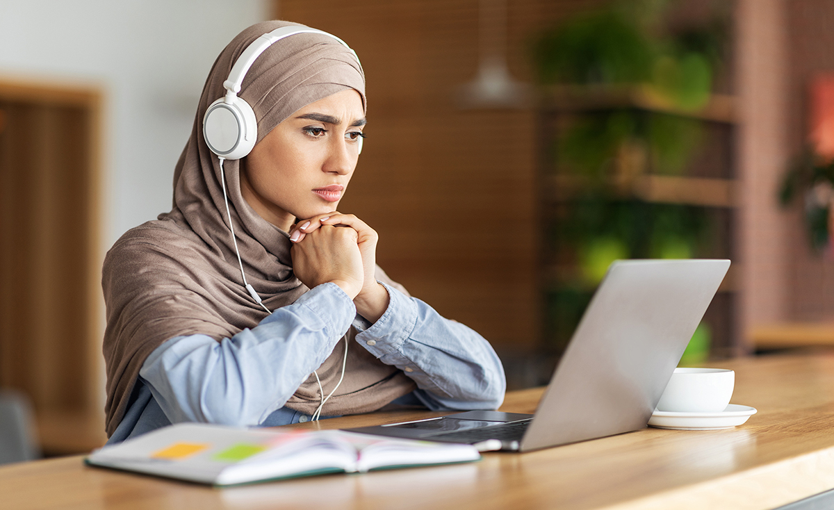 Photo of a young woman wearing a hijab and headphones watching her laptop screen intently with her hands clasped.