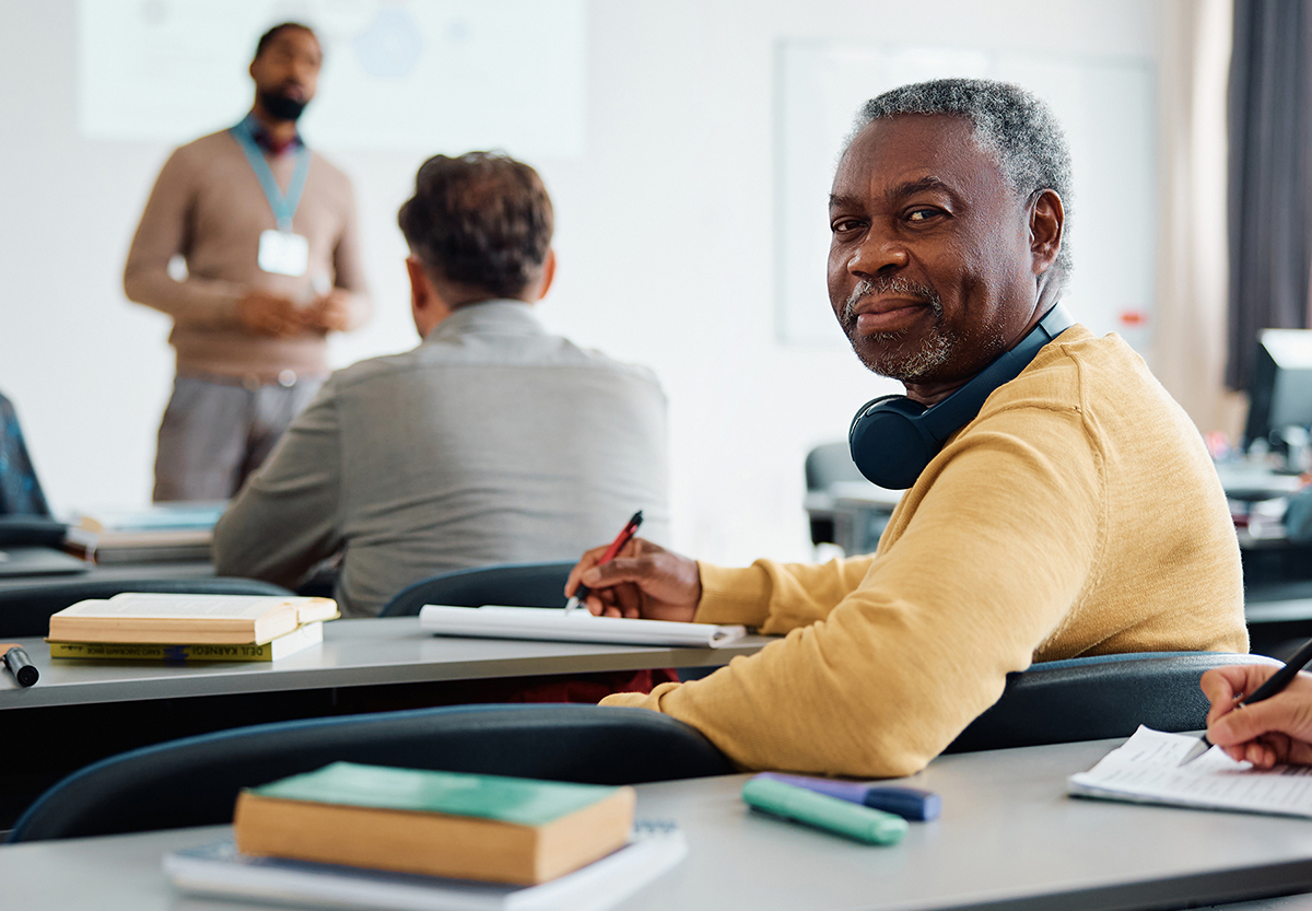 Photo of an older man seated at a desk in a classroom with other adult student with a teacher standing at the front of the class in the background. 