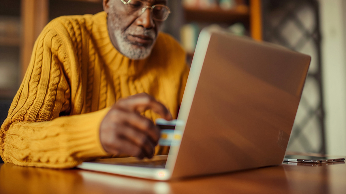 Photo of an older African American man seated in front of a laptop holding a credit card.