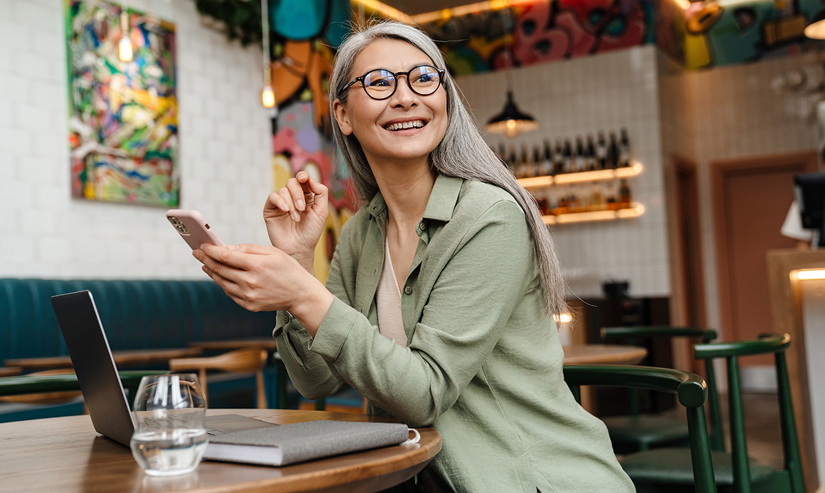 Photo of a smiling older woman sitting in a coffee shop holding a smartphone.