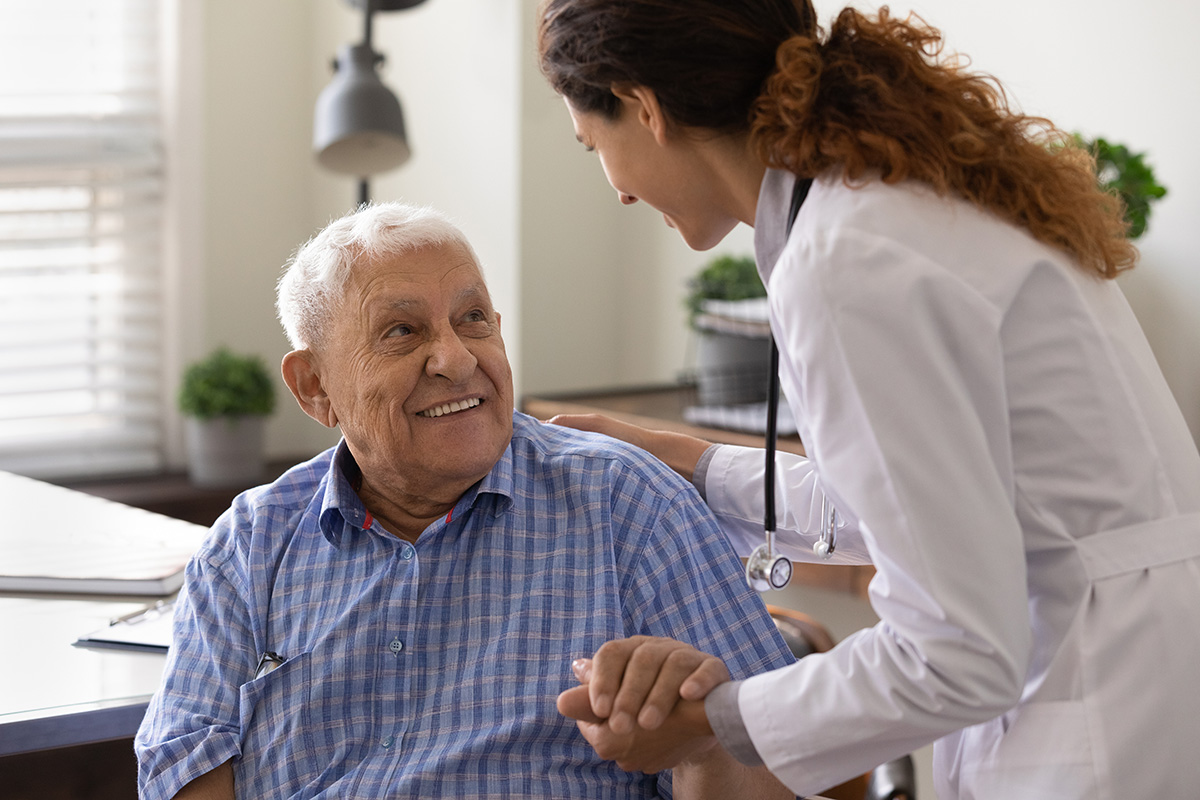 Photo of an older man smiling at a young woman dressed in a white lab coat wearing a stethoscope. 