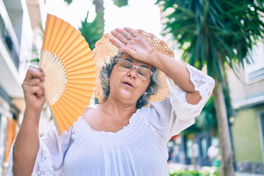Photo of an older woman outdoors on a hot day wearing a hat and holding a fan while wiping her forehead.